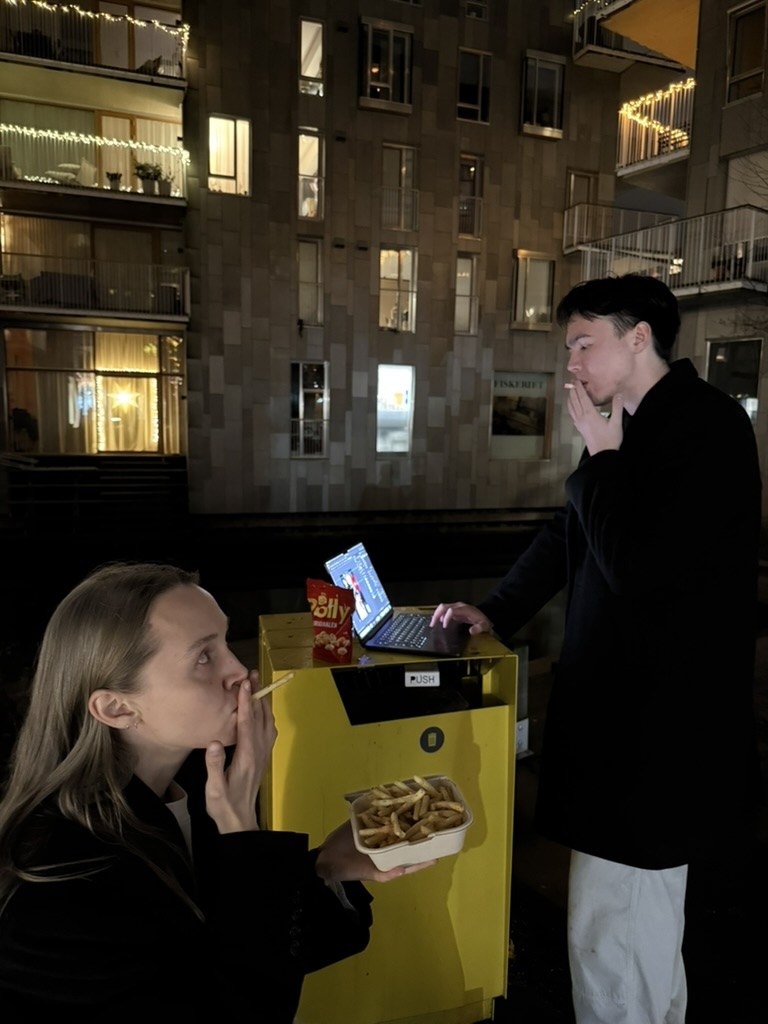 Two people at night beside a yellow deposit bin used as a standing desk, with a laptop and a tray of chips on top
